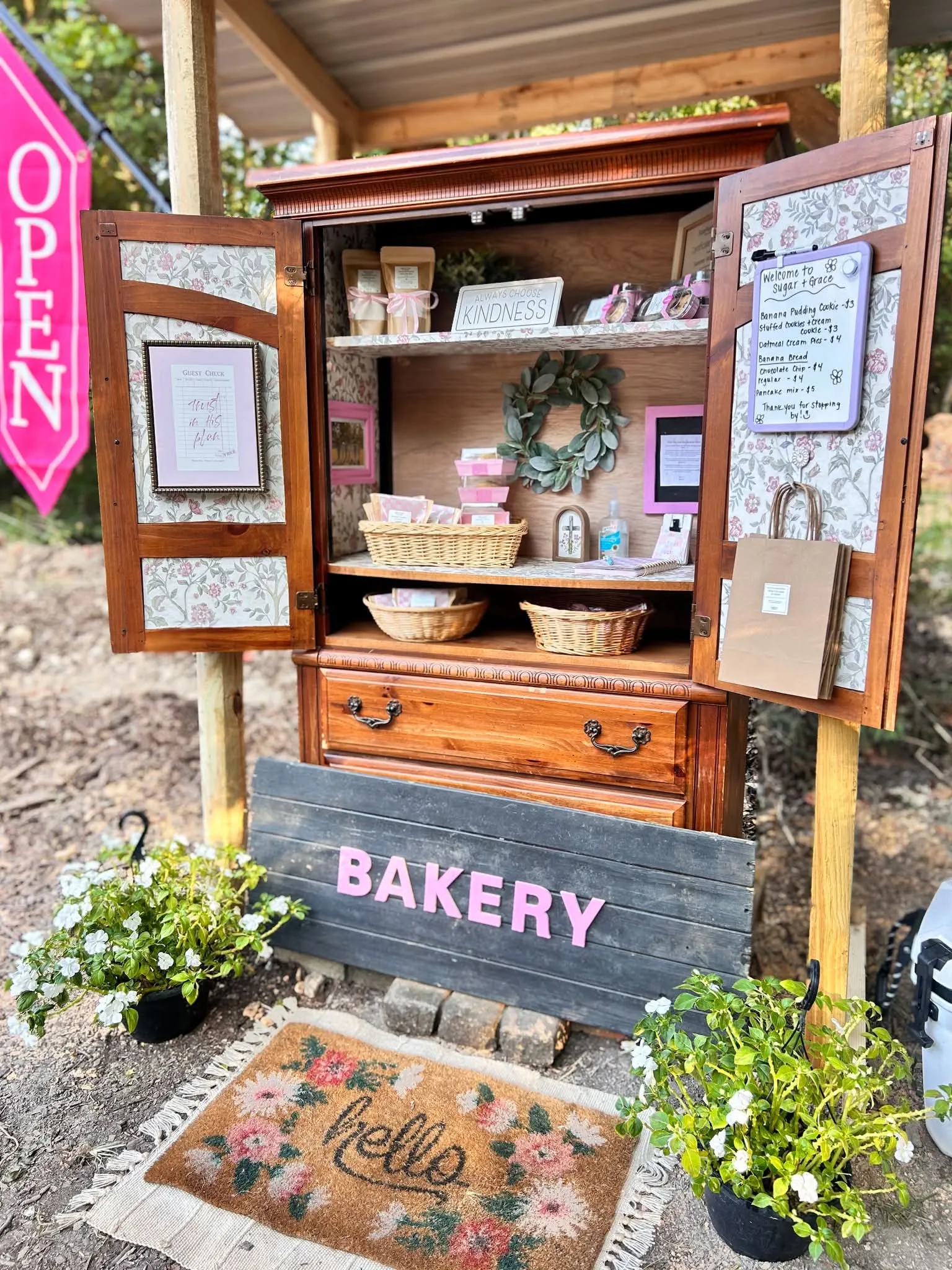 The Sugar & Grace stand, a repurposed wooden armoire with its doors open, shelves stocked with cookies, banana bread and jarred treats beside a hand-lettered Bakery sign
